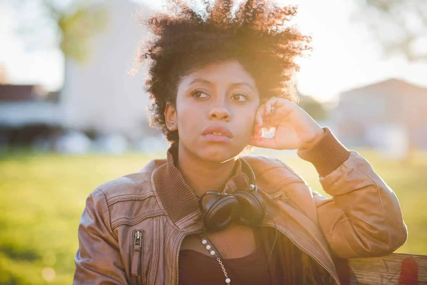 Black girl wearing brown leather jacket with curly hair, standing outside in the sunlight leaning on a fence post. 