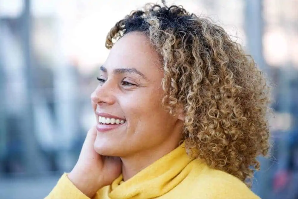 A beautiful African American woman with blonde, brown curly hair strands styled with coconut oil and organic hair oil.