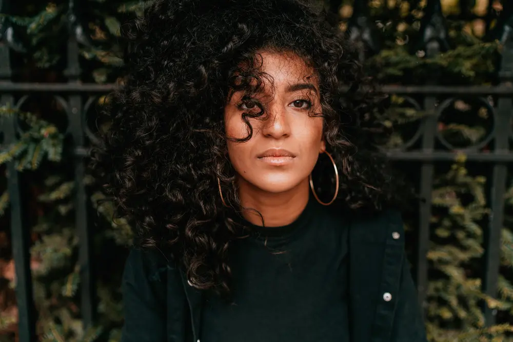 Women sitting on a park bench with sunflower oil treated hair wearing bronze earrings, curly hair and a green jacket. 