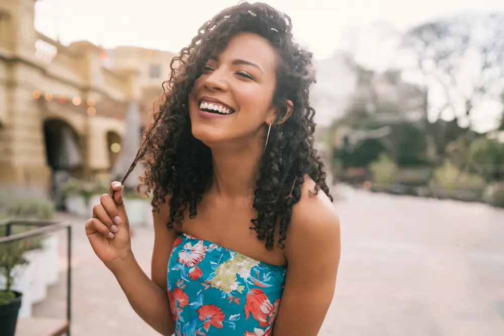 Beautiful Latin confident woman pulling a hair strand covered in a few drops of almond oil laughing in the city street.