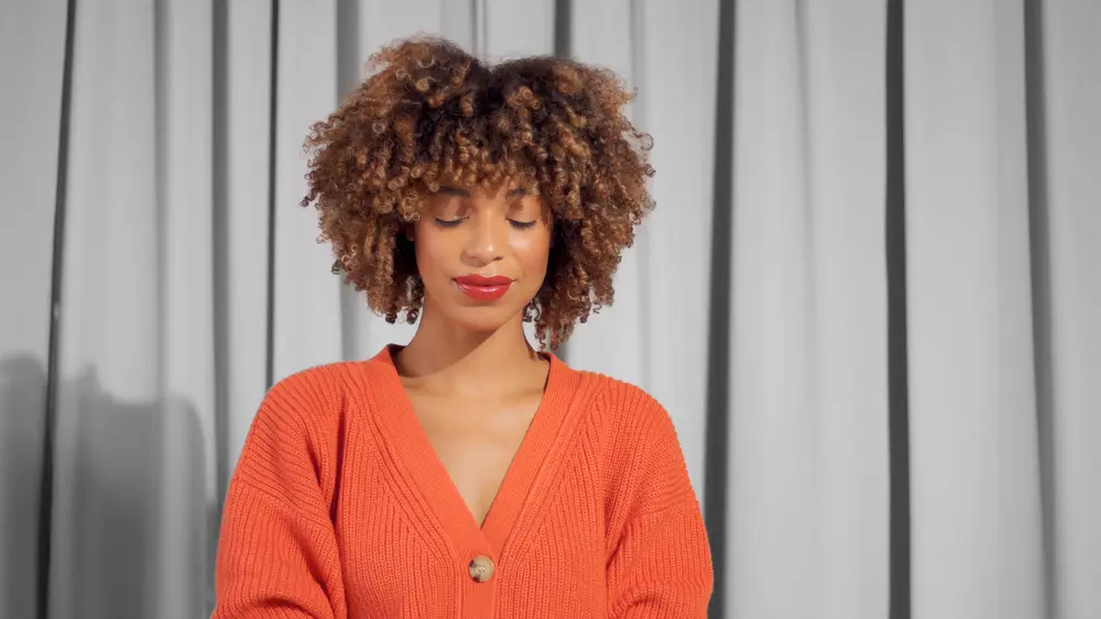 Cute African American female with a tight textured 3b curly afro hair in bright orange jacket with natural makeup for dark skin tones looking down at the ground.