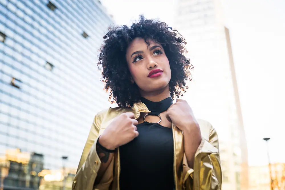 Black girl wearing a gold jacket and black dress with curly type 3b hair and red lipstick.