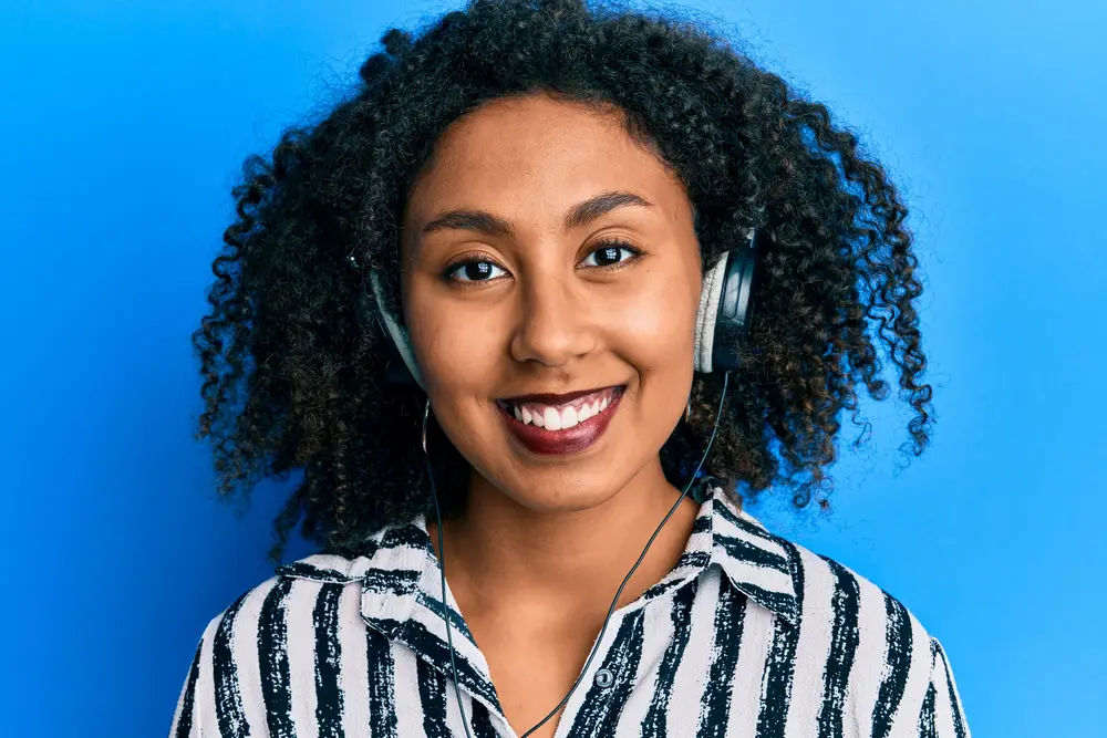 Beautiful African American woman with afro hair wearing call center agent headset looking positive and happy standing and smiling with a confident smile showing teeth.