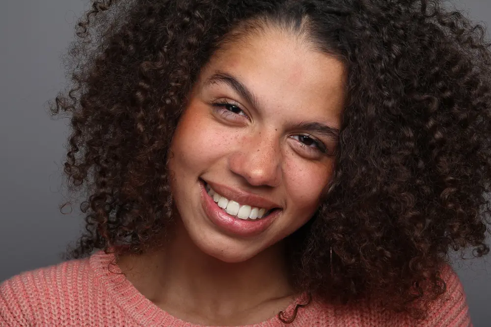 Black woman with freckles, curly hair and a big smile wearing a pink sweater looking directly into the camera.