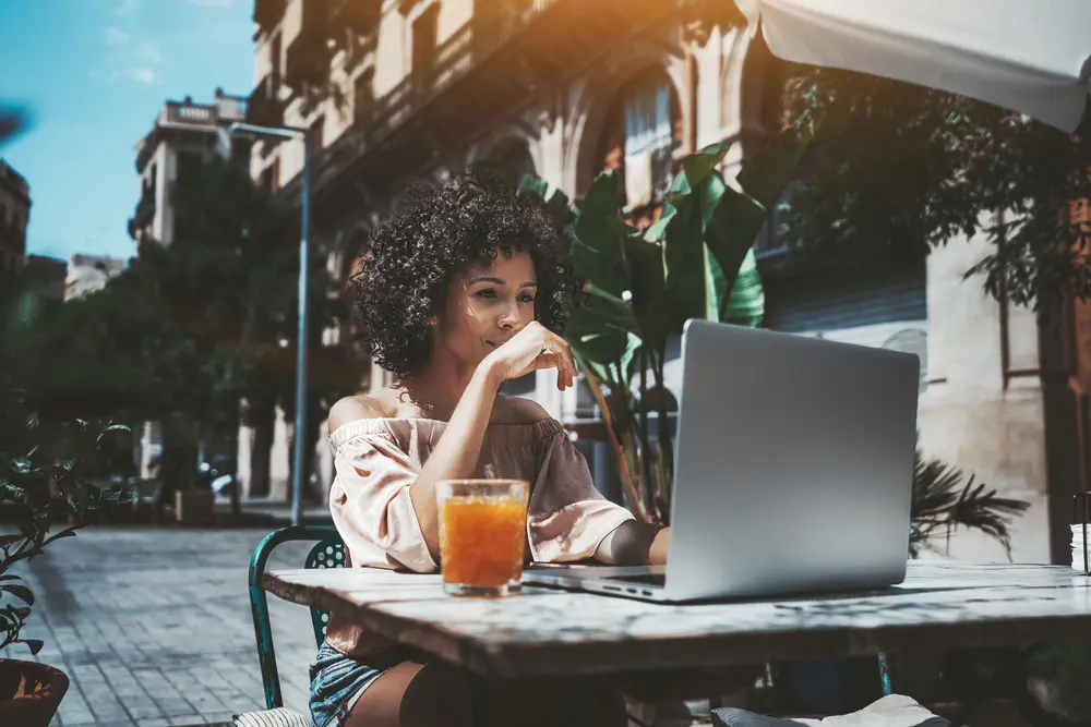 An attractive biracial girl with natural curls takes a photo with her iPhone while sitting at an outdoor bar on a sunny summer day wearing an orange shirt and blue jean shorts. 