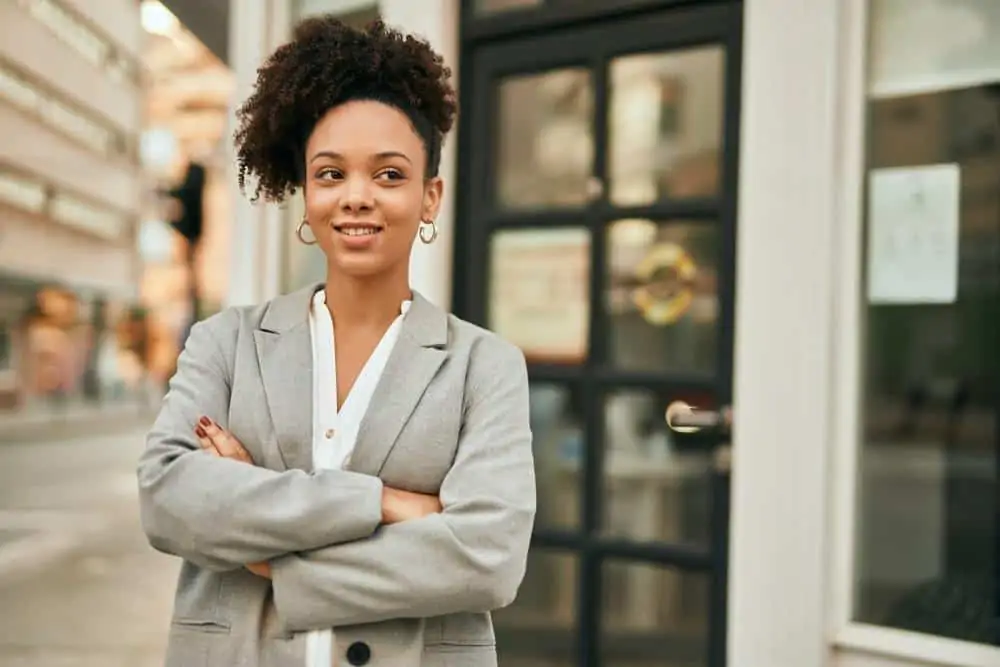 Young African American businesswoman with arms crossed standing at the city, smiling with gold ear rings, and a curly pineapple hairdo that's been treated with peppermint oil.