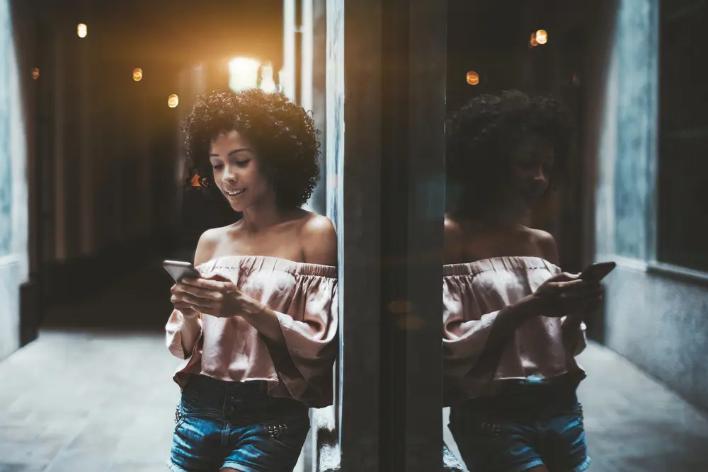 A cheerful young Brazilian female with a medium curly hairstyle is leaning against the wall on a partly dark street corner.