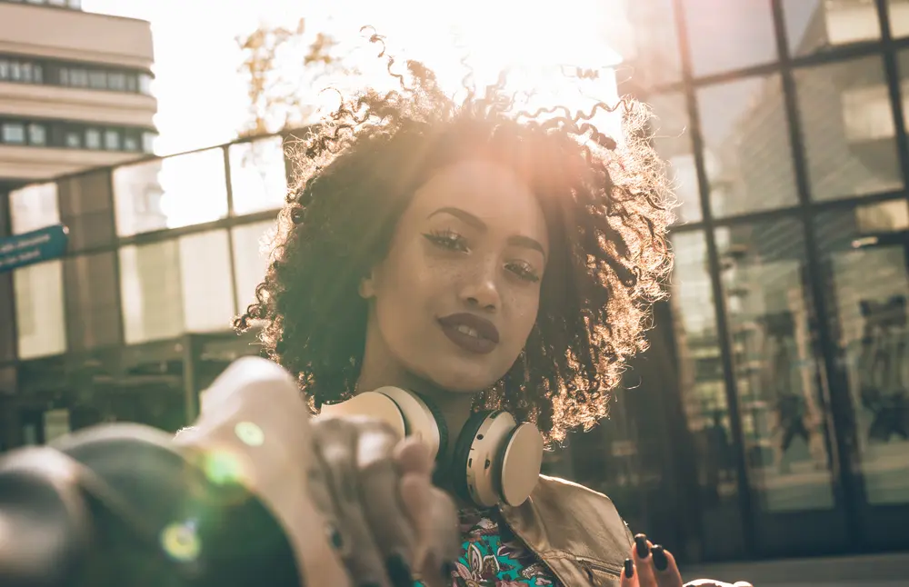 Latin woman with curly hair that scented with cedarwood oil  standing on a street corner wearing  beats headphones.