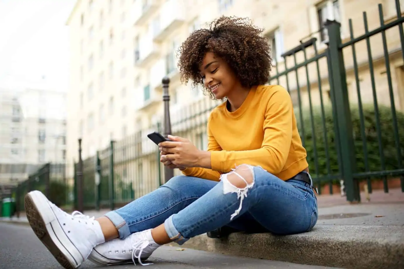 Beautiful African American female with curly hair sitting on a curb using her mobile phone wearing jeans, a yellow shirt and converse. 