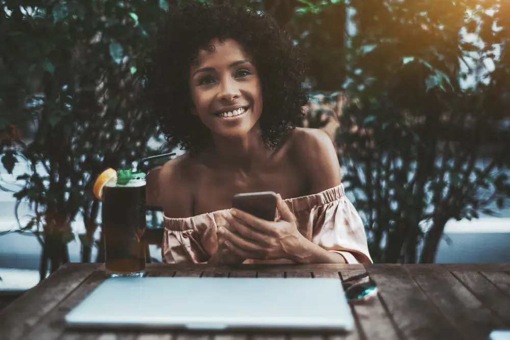 A laughing young curly haired Brazilian female with the smartphone, notebook and a cocktail in a street bar.