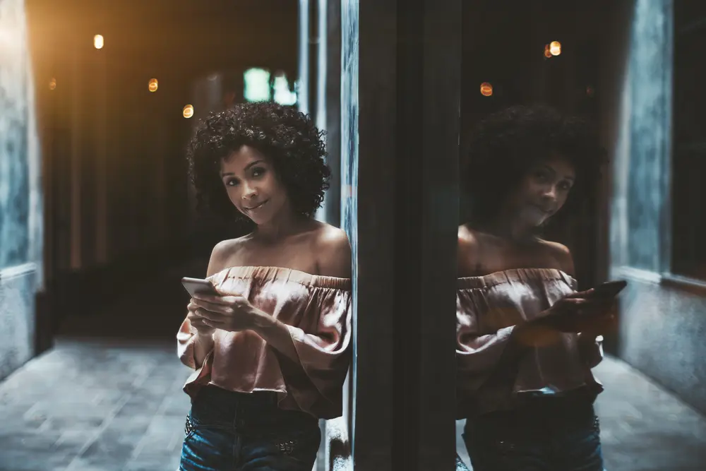 A cheerful young African-American female with the cellphone in her hands is leaning on the wall with a mirror in front which reflects her image.