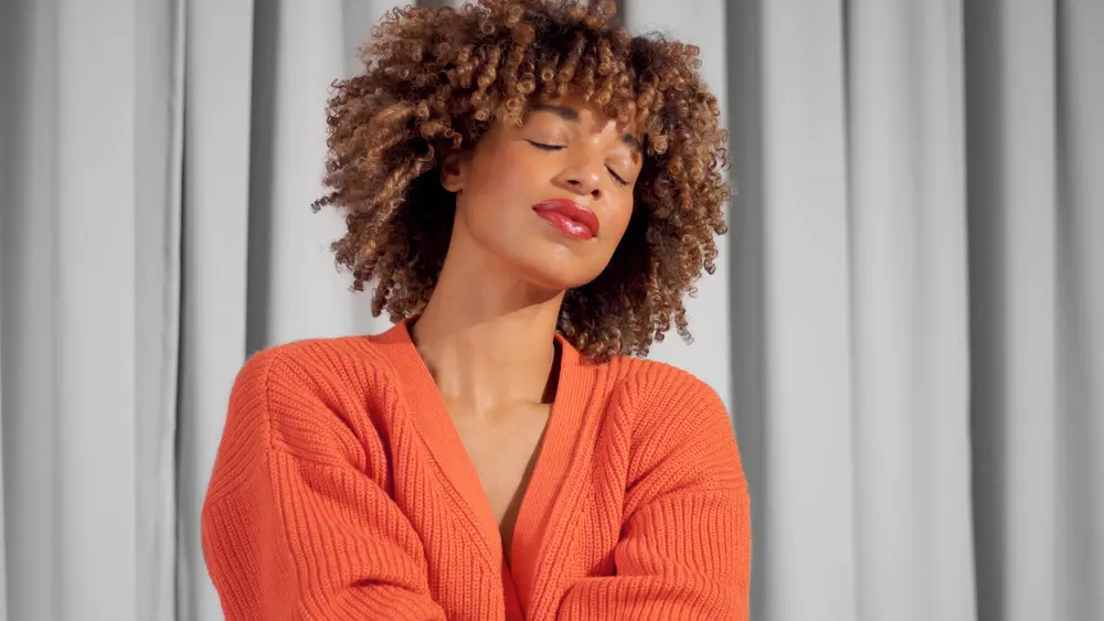 Mixed race black woman with Shirley Temple curls with rollers wearing a bright orange jacket with natural makeup for dark skin tones with her eyes closed.