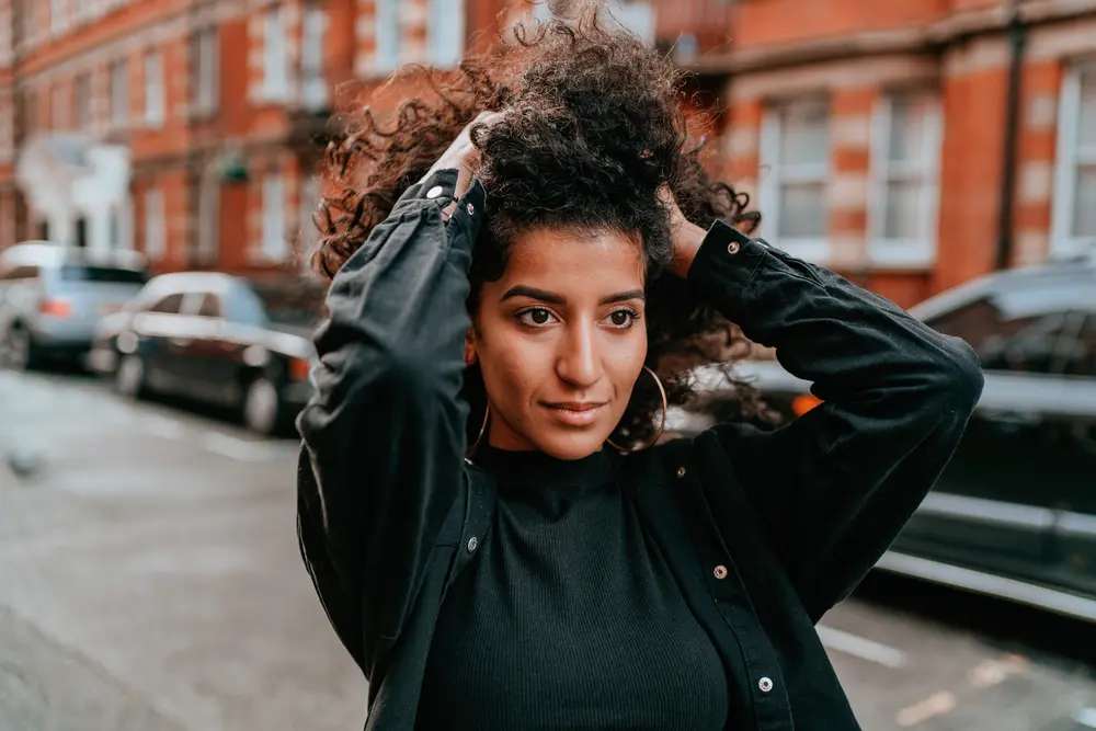 Portrait of charming young woman with curly hair walking down the street with cars in the background.