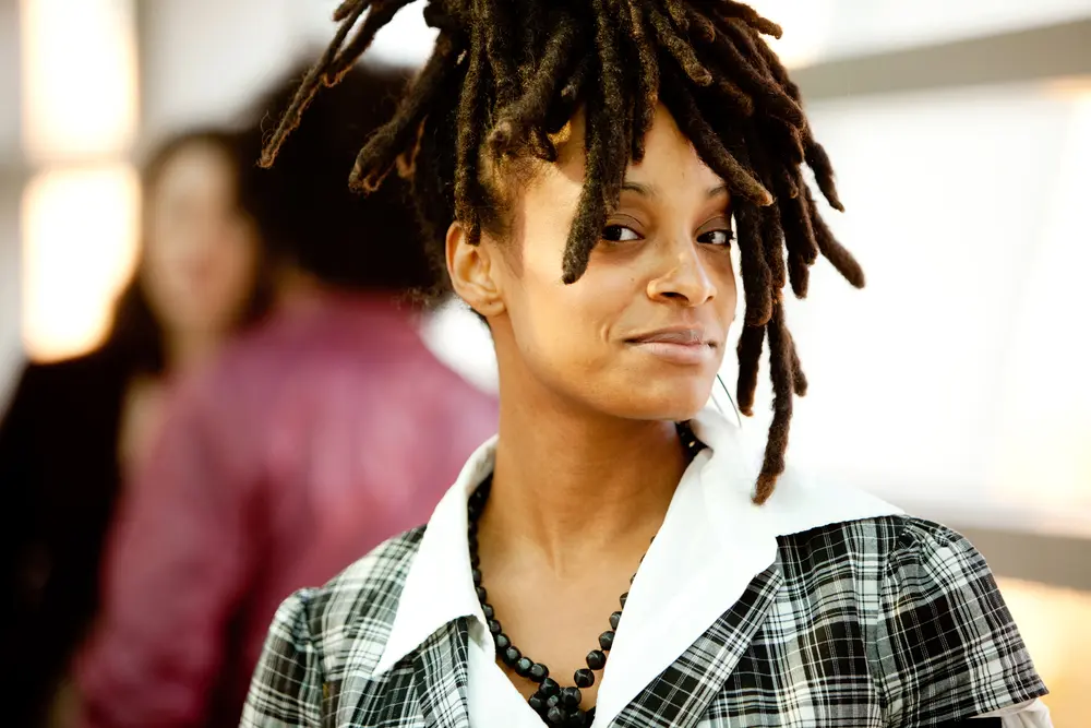 Young African American female with a green and white plaid shirt, with a white collard undershirt and black beaded necklace looking directly into the camera. 