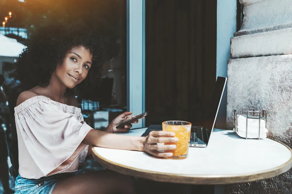 Charming young curly hair Brazilian woman in a street cafe with the cellphone, netbook, and the glass of orange juice.