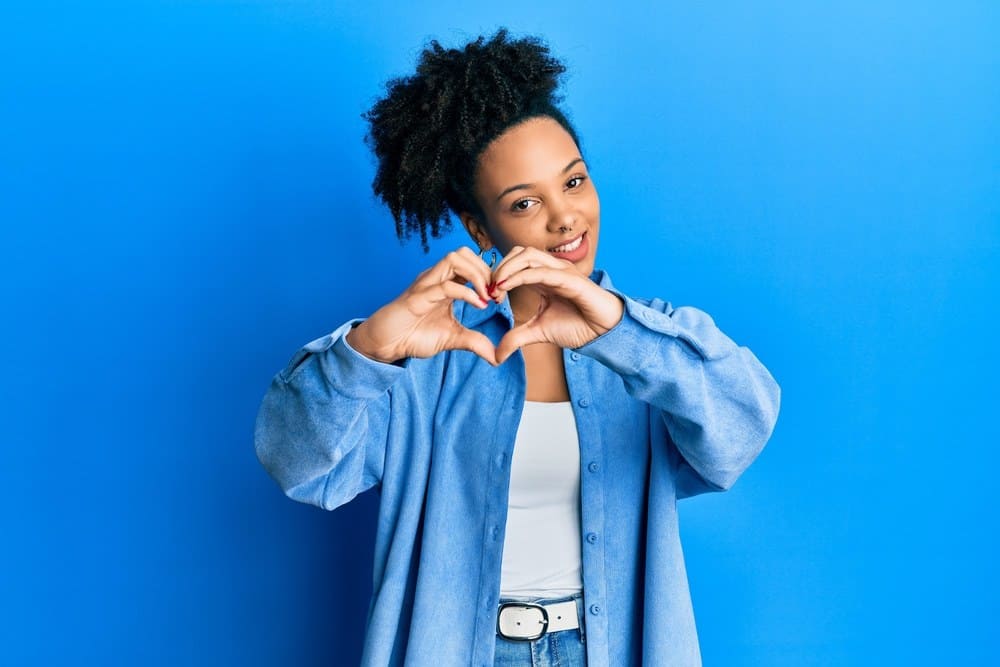 Young African American girl wearing casual clothes smiling in love doing heart symbol shape with hands.