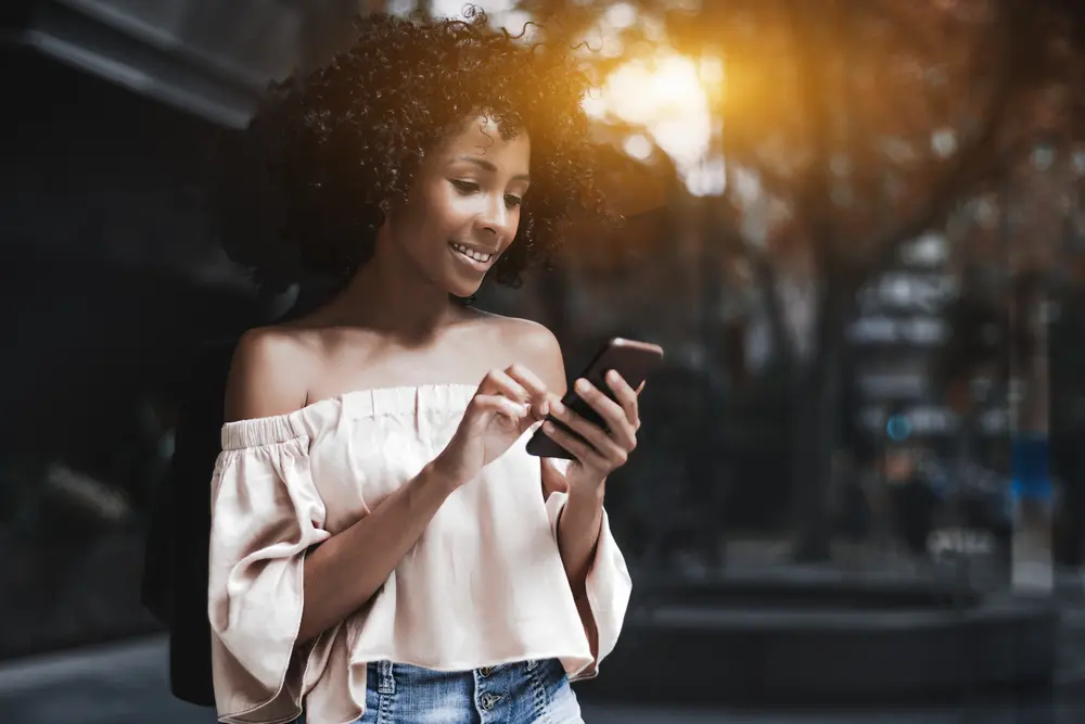 African-American female with naturally curly heatless curls carrying an Android cellphone on a summer city street typing a message while standing outdoors near a glass wall.