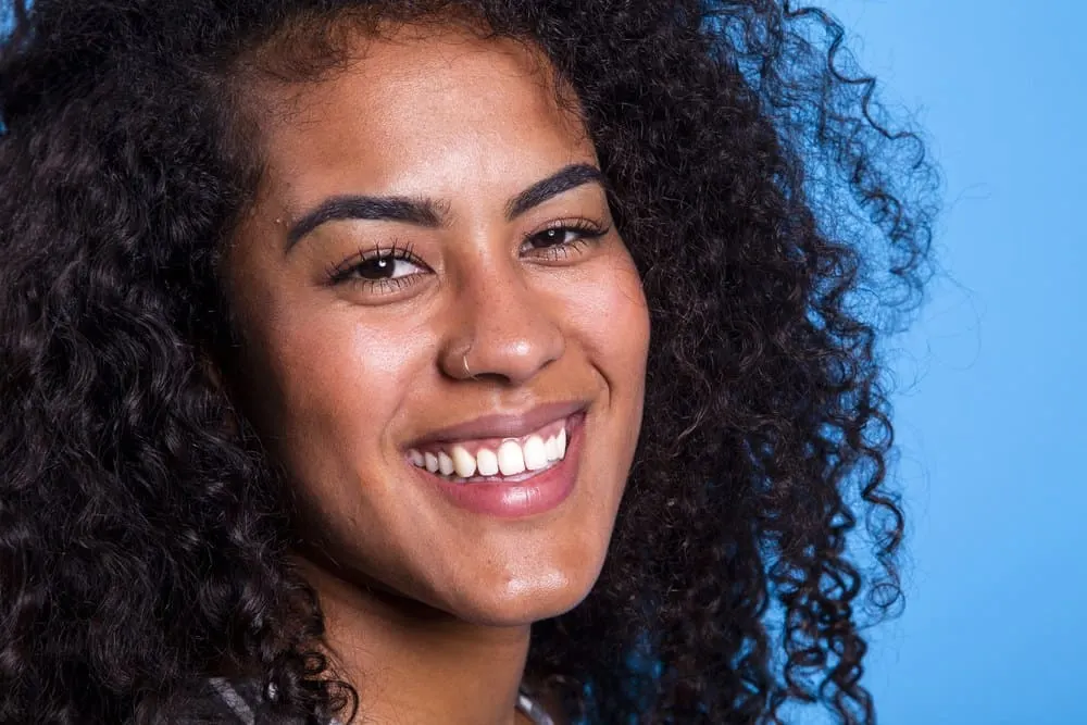 Does Ayurveda oil work is that question that this young woman wonders as she smiles against blue background with naturally curly type 2 hair. 