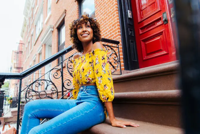 Black woman sitting outside an apartment building on stairs wearing a yellow shirt and blue jeans