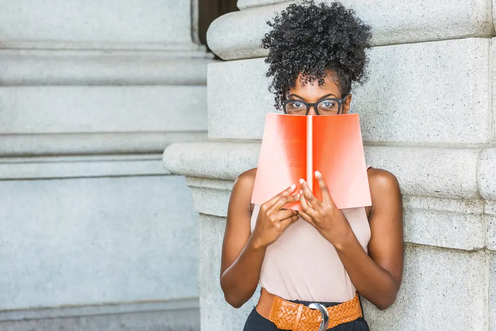 Pretty black girl wearing a pink shirt, brown belt while reading a book outside the library. She's wearing black glasses and has naturally curly type 4A hair.