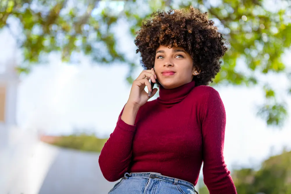 Young black woman speaking on mobile phone, while sitting on a park bench wearing a red turtle neck sweater. 