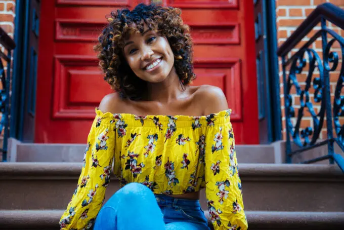 Light skin black girl sitting outside an apartment building on the stairs wearing a yellow shirt and blue jeans.