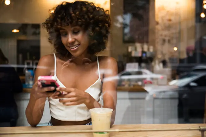 Black lady wearing a white spaghetti strap dress sitting in a coffee shop drinking a latte while using her mobile phone