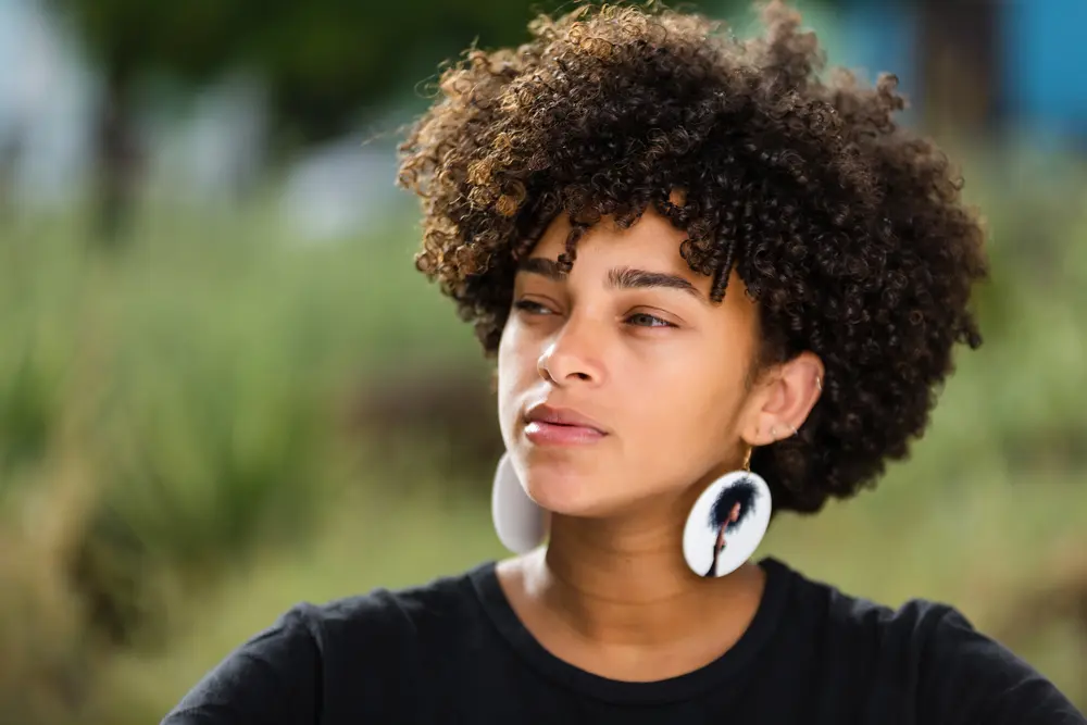 Young African American woman with curly hair strands wondering what is low porosity hair while standing outside.