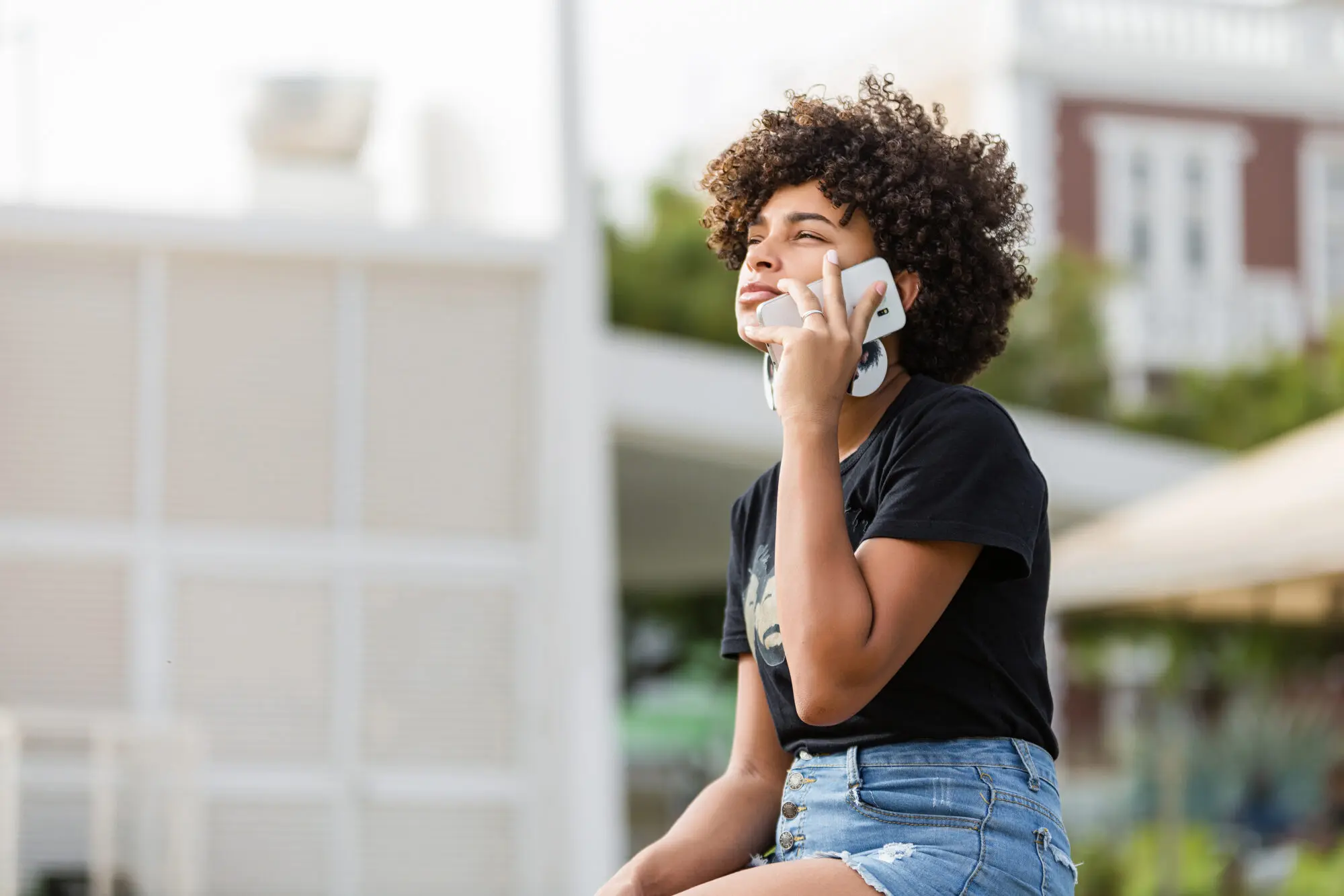 Women wearing cut-off blue jean shorts and a black t-shirts with African American images, sitting on a park bench while using the phone. 