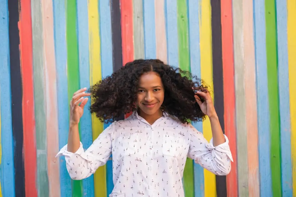 Black women with long natural hair standing in front of a multi-colored wooden wall admiring her beautiful curly hair. 