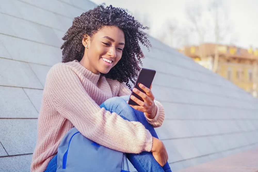 Black girl reading beauty tips about finger coil styles while using her mobile phone and sitting on a rooftop. 