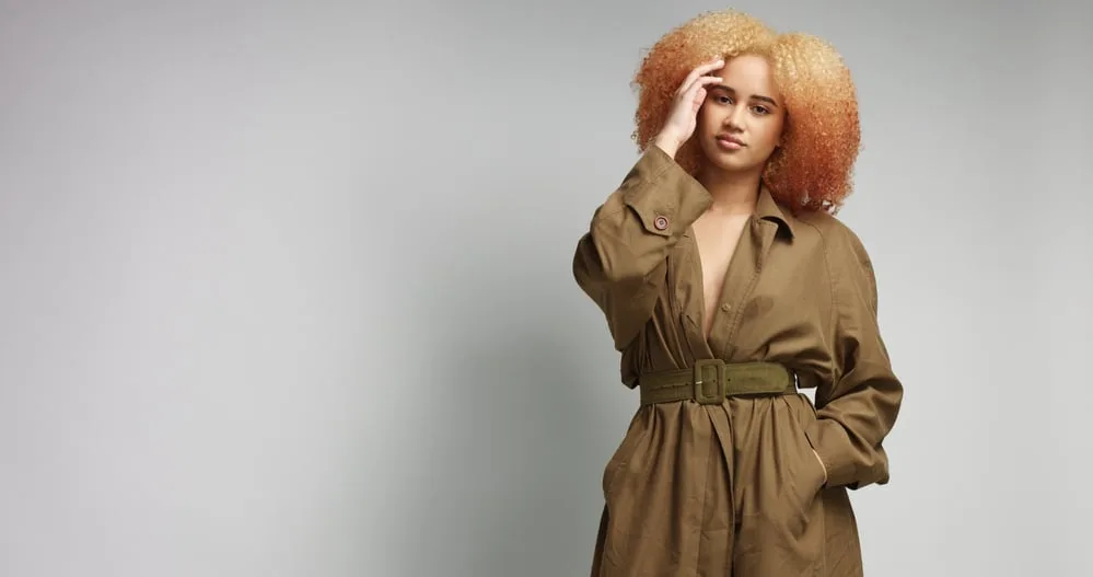 Black women wearing during a photoshoot wearing a khaki coat standing in front of a gray wall