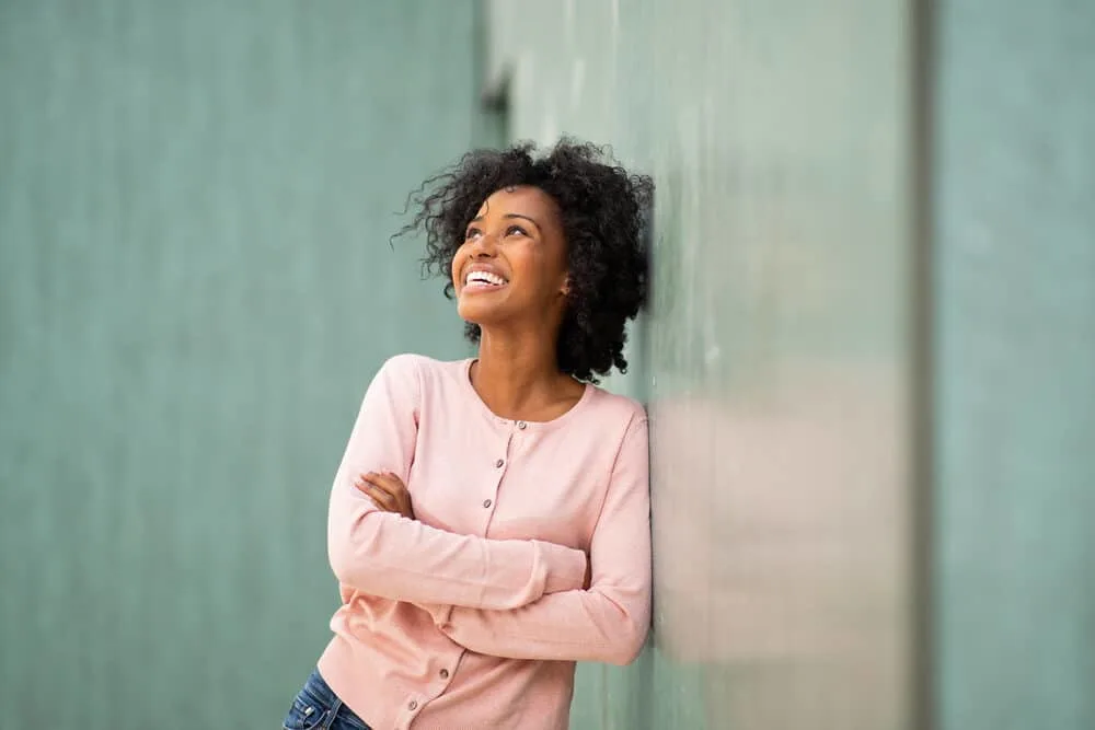 African American female leaning against a green marble wall wearing a pink shirt and casual pants. 