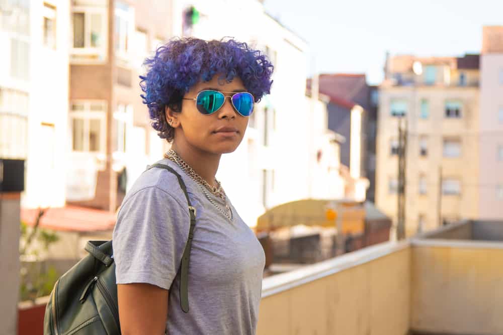  Femme latine debout à l'extérieur portant un t-shirt gris, des chaînes de cou et un sac à dos vert.