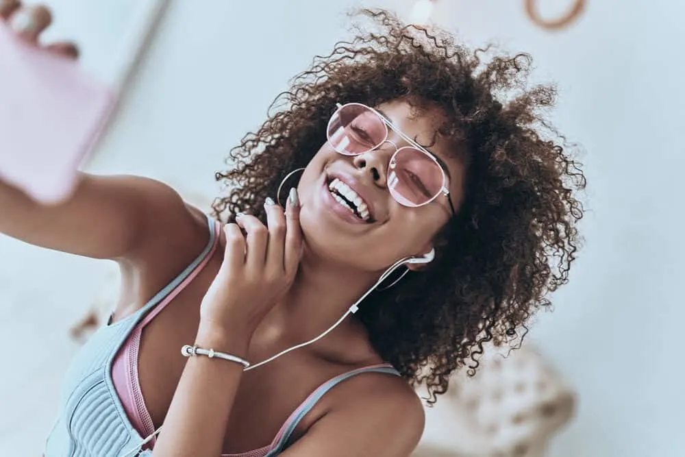 Female with pink-tinted glasses, Apple earphones, blue nail polish, and pink lipstick.