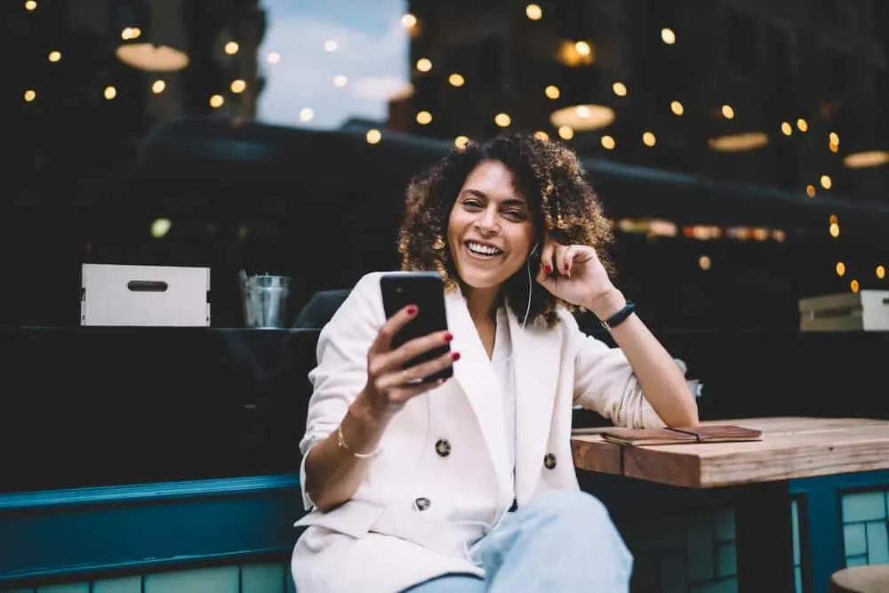 African American female with naturally curly hair wearing a pink coat, white shirt, and blue jeans.