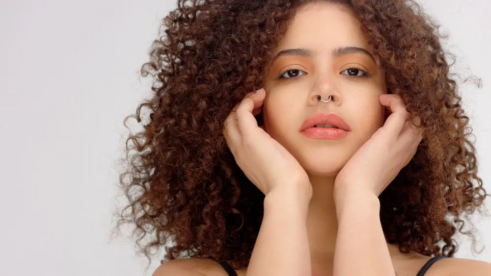 Hispanic girl with her hands on her face with wavy hair starring directly into the camera. 