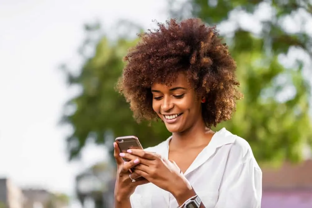 Black girl leaving the hairdresser wearing a white shirt, pink nail polish, and light brown hair dye.