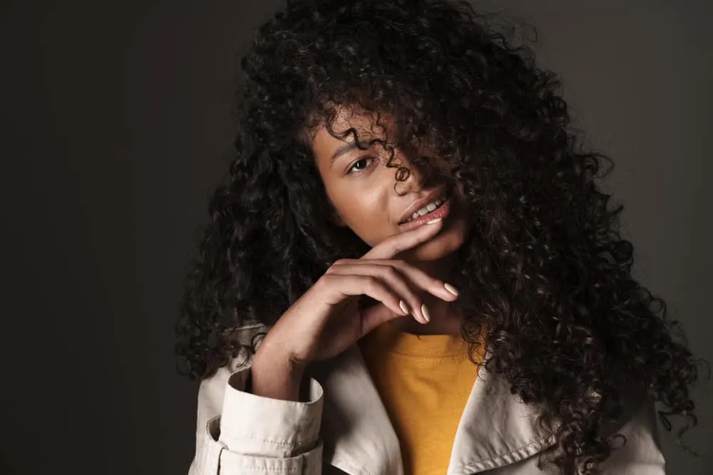 Black female wearing a beige jacket and orange undershirt with very wavy hair.