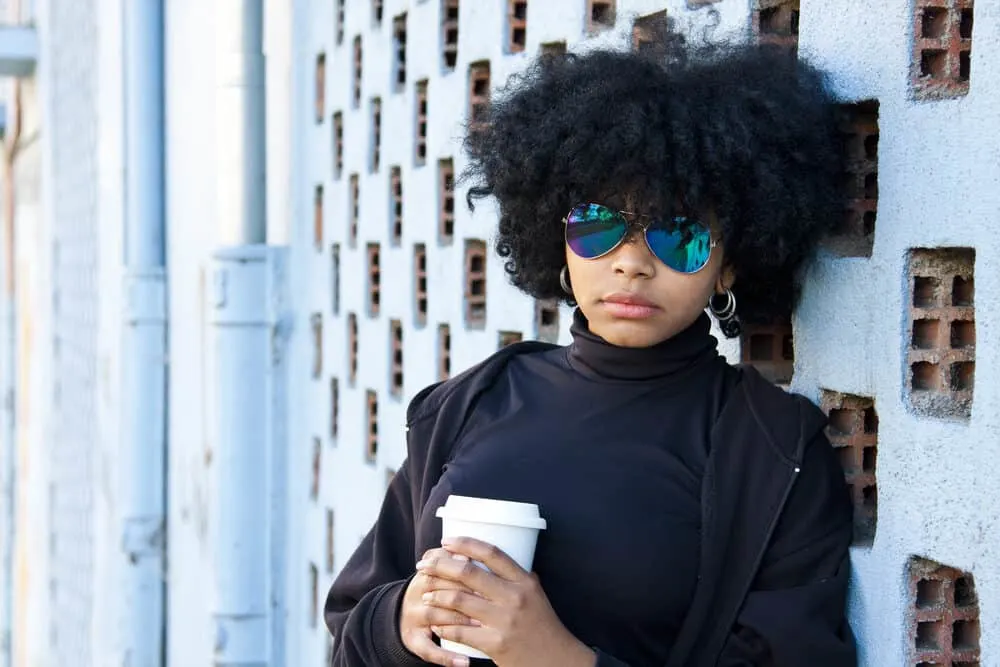 Lady standing outside drinking coffee wearing a mock turtleneck and hoop earrings.