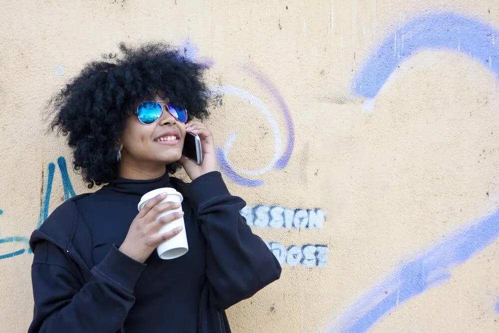 Black women with curly hair wearing shades while drinking a cup of coffee outside.