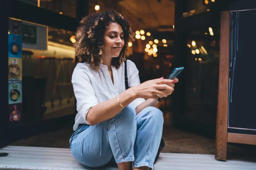 Happy female sitting on the floor in a cafe using her cell phone with ombre curly hair strands.