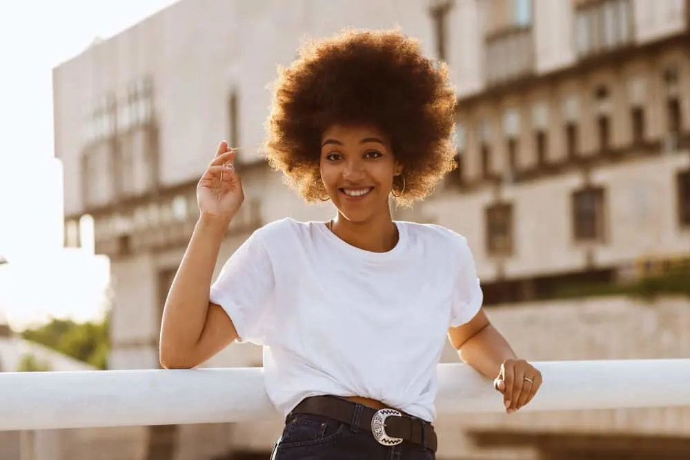 Female with 4c hair strands with a blonde tone provided by the sunlight wearing a white t-shirt leaning against a fence