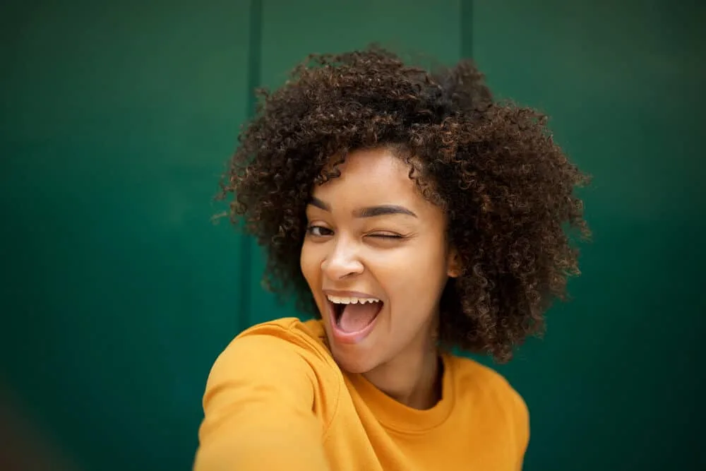 Black girl winking her left eye with a hair grease mix in her naturally curly hair strands.