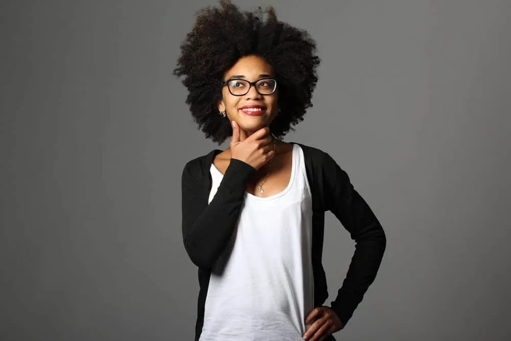 A black woman with type 4c hair holding her chin wearing a white T-shirt, black glasses, earrings, and red lipstick.