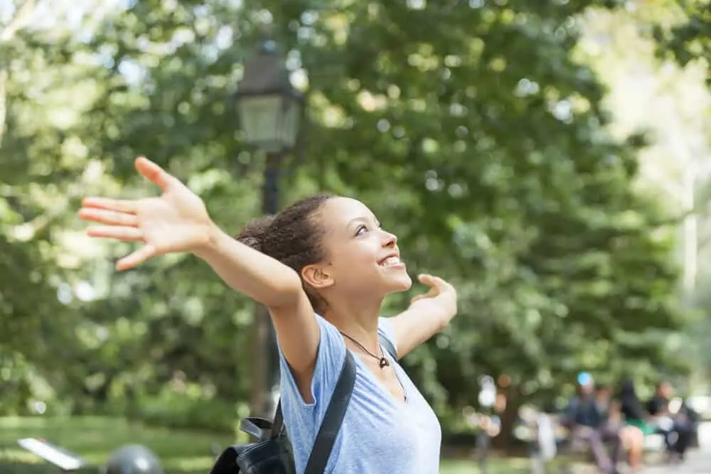 A cute young black female with head lice has her arms stretched out towards the sky wearing a gray t-shirt.