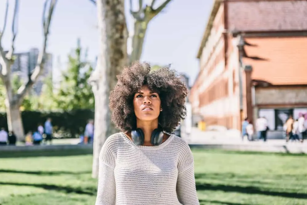 A woman with her own hair dyed with brown highlights walking through the park with headphones.