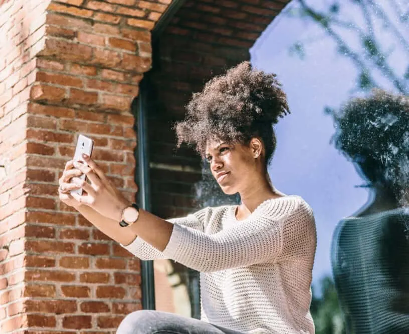 Beautiful young African American girl with a joyful expression, taking a selfie next to a large window and looking at herself in the reflection.