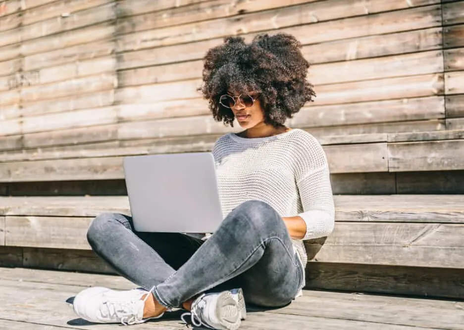 A young stylish woman wearing tape hair extensions sitting in a park and working on her laptop while having one leg crossed over the other.