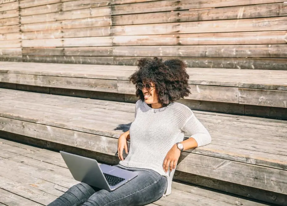 Beautiful young woman wearing an afro wig blending perfectly with her own hair while sitting on a street and smiling while working on her laptop.